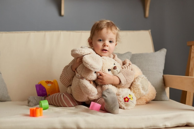 Poziome ujęcie uroczego blond niemowlęcia siedzącego na kaszlu z miękkimi zabawkami obejmującymi królika i misia patrzącego w kamerę spędzającego wolny czas w domu
