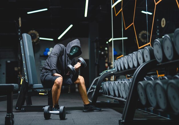 Poziome zdjęcie silnego mężczyzny w masce siedzącego na ławce, pozującego w nowoczesnej hali sportowej Nierozpoznawalny mężczyzna patrzący w kamerę