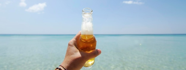 Poziomy baner lub nagłówek z męską ręką trzymającą butelkę piwa na tle słonecznego nieba i krystalicznie czystego morza Koncepcja wakacji