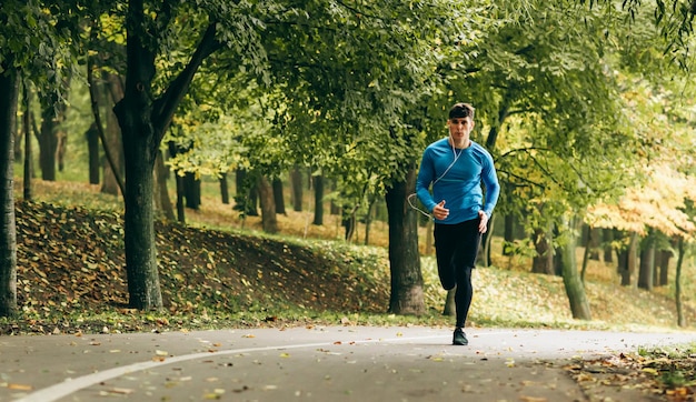 Poziomy obraz całego ciała młodego joggera joggingu na świeżym powietrzu w tle lasu Fitness mężczyzna ćwiczących w parku na sobie niebieską i czarną odzież sportową Ludzie i koncepcja sportu