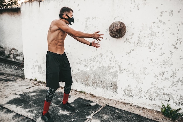 Poziomy obraz muskularny mężczyzna robi ćwiczenia z piłką lekarską w masce na betonowym tle na zewnątrz z miejscem na reklamę Półnagi sportowiec robi trening