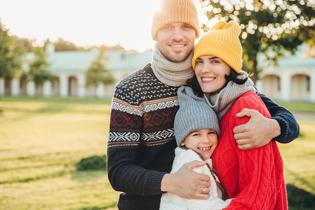 Zdjęcie poziomy portret młodego przystojnego mężczyzny w żółtym kapeluszu i ciepłym swetrze z dzianiny obejmuje żonę i córkę pozuje do kamery, stojąc w parku trzech członków rodziny spędza razem weekendy