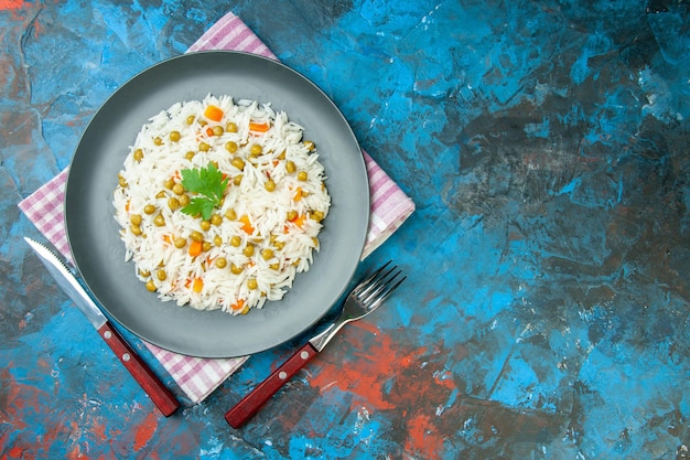 Zdjęcie poziomy widok pysznego posiłku ryżowego z marchewką pisum serwowaną z zielenią na talerzu na fioletowych sztućcach z ręczników w paski po prawej stronie na mieszanym kolorowym tle