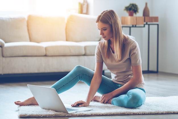 Pozostań w kontakcie w domu. Piękna młoda kobieta korzysta z laptopa siedząc na dywanie w domu