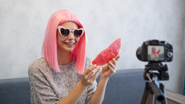 Pozytywna blogerka nagrywa film o zdrowym odżywianiu i trzyma w dłoniach arbuza. Kobieta ubrana w różową perukę i okulary w kształcie serca