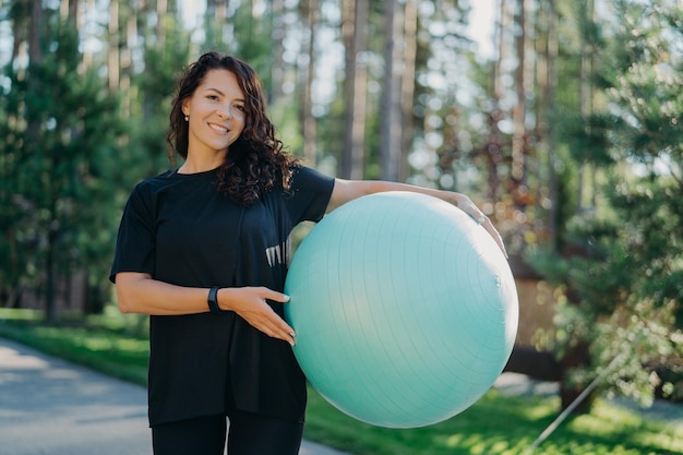 Pozytywna brunetka prowadzi aktywny tryb życia trzyma piłkę fitness na tle lasu ubrana w czarne ubrania ma szczęśliwy uśmiech Ładna kobieta przygotowuje się do treningu pilates robi ćwiczenia