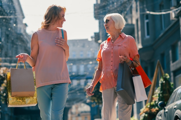 Pozytywna dorosła kobieta i starsza pani uśmiechają się do siebie na ulicy podczas spaceru z papierowymi torbami na zakupy