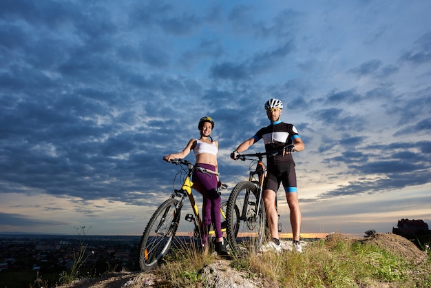 Pozytywna dziewczyna i mężczyzna pozuje z rowerami w hełmach i odzieży sportowej.