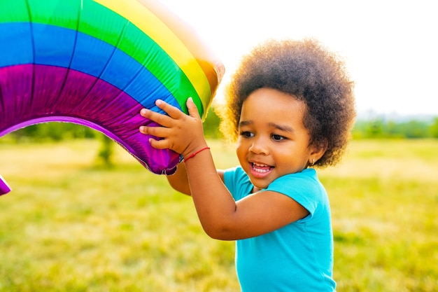 Pozytywna dziewczynka rasy mieszanej z afro kręconymi włosami trzymająca tęczowy balon w letnim parku