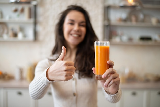 Pozytywna kaukaska dama trzymająca szklankę organicznego soku pomarańczowego i pokazująca kciuk stojący w jasnym wnętrzu kuchni