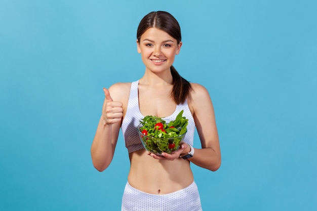Pozytywna kobieta pokazująca kciuki do góry trzymająca dużą miskę z sałatką ze świeżych warzyw, utrzymująca dietę detoksykacyjną