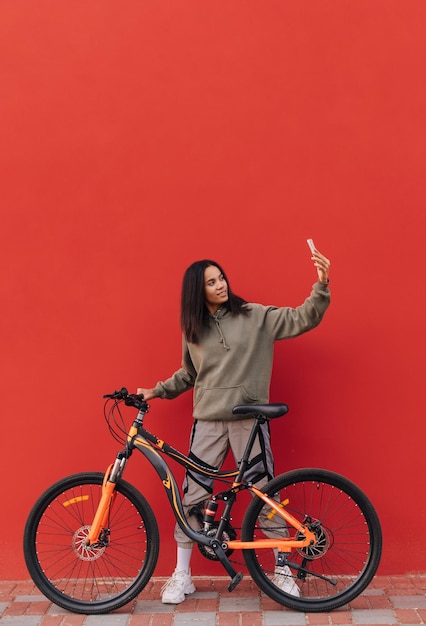 Pozytywna Kobieta Siedząca Na Rowerze Na Tle Czerwonej ściany I Robiąca Selfie