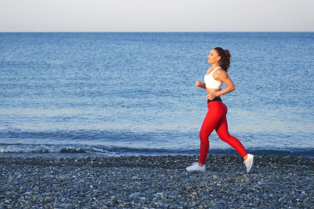 Pozytywna kobieta sprawny na letnim porannym joggingu na plaży w czerwonych legginsach na tle wybrzeża morskiego