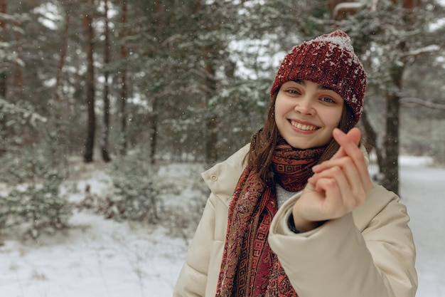 Pozytywna kobieta w ciepłych ubraniach pokazująca gest serca lub miłości palcami w śnieżnym lesie