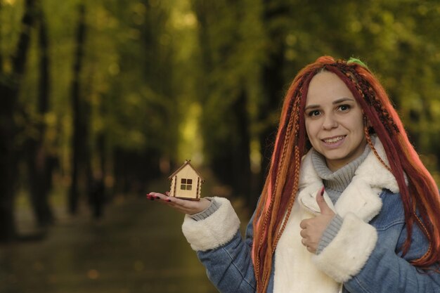 Pozytywna kobieta z dredami trzymająca mały drewniany domek w parku leśnym Kobieta z domkiem zabawek patrząca na kamerę uśmiechnięta Koncepcja zakupu nowego dachu nad głową w ekologicznym miejscu