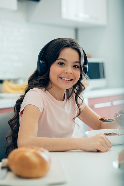 Pozytywna ładna dziewczyna siedzi przy stole z łyżką w dłoni i podczas jedzenia słucha muzyki w słuchawkach