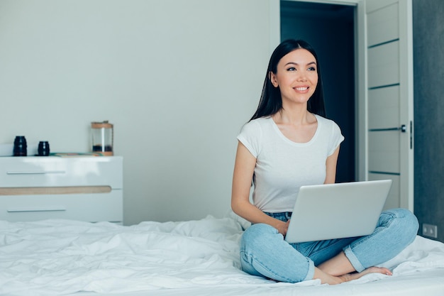 Pozytywna ładna pani ciesząca się odległą pracą i uśmiechnięta siedząc na łóżku z nowoczesnym laptopem Baner strony internetowej