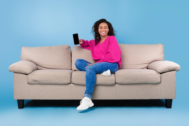 Pozytywna młoda czarna dama siedzi na kanapie z telefonem komórkowym na niebieskim tle studia makieta dla aplikacji
