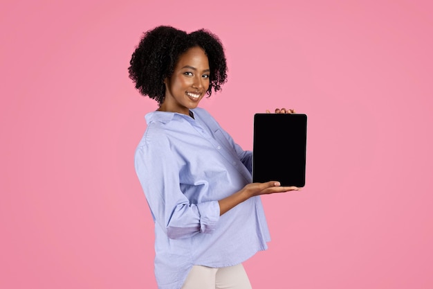 Pozytywna młoda czarna kobieta w ciąży w zwykłym tablecie z pustym ekranem poleca aplikację