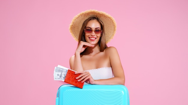 Pozytywna młoda dziewczyna w słomkowym kapeluszu i okularach przeciwsłonecznych z biletami i walizką różowe studio tło uśmiech...