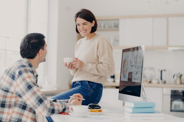 Pozytywna młoda kobieta i mężczyzna omawiają wynajmowanie mieszkania w przestrzeni coworkingowej z komputerowymi i papierowymi dokumentami szczęśliwa żona pije kawę wygląda z przyjemnym uśmiechem u męża zarządza budżetem