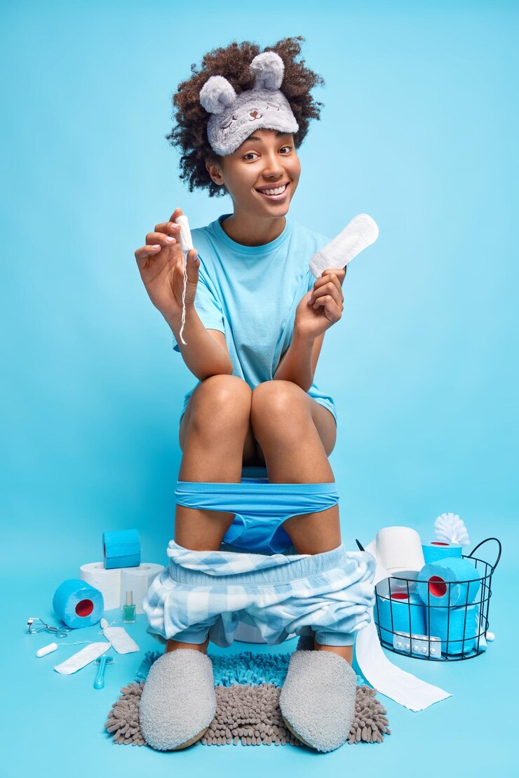 Pozytywna młoda kobieta ma menstruację trzyma tampon i podpaski na muszli klozetowej, ubrana w zwykłe ubrania pozy w toalecie na białym tle nad niebieskim tłem. Koncepcja higieny kobiet