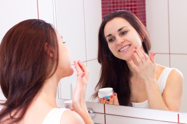 Pozytywna młoda kobieta o długich ciemnych włosach w białej koszuli, używająca kremu do twarzy przed lustrem w swojej łazience