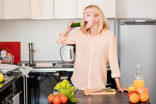 Pozytywna młoda kobieta pozuje w kuchni w domu. Głodna blondynka na sobie dorywczo pasiastą koszulę, jedzenie ogórka podczas przygotowywania sałatki jarzynowej, patrząc na kamery. Zdrowa żywność, koncepcja stylu życia.