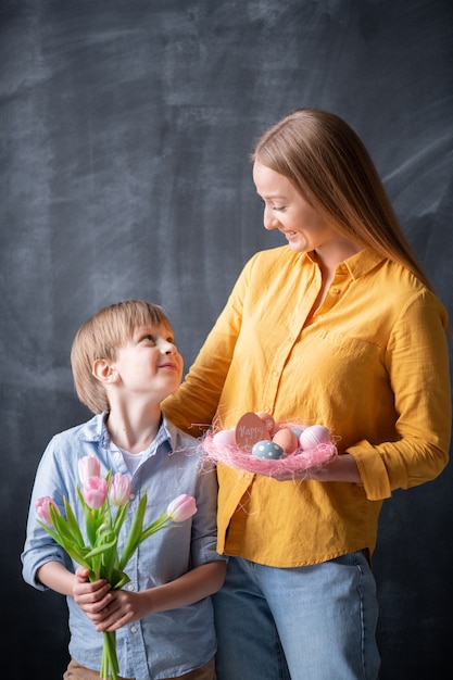 Pozytywna Młoda Mama Z Wielkanocnym Gniazdkiem I Jej Syn Z Bukietem Tulipanów, Patrząc Na Siebie Podczas Zabawy Na Wielkanocnych Wakacjach