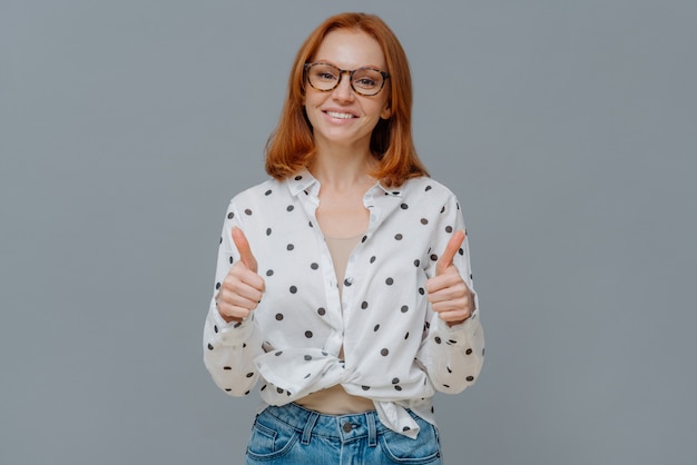 Pozytywna rudowłosa kobieta o łagodnym uśmiechu pokazuje kciuki do góry, demonstruje gest lub aprobatę