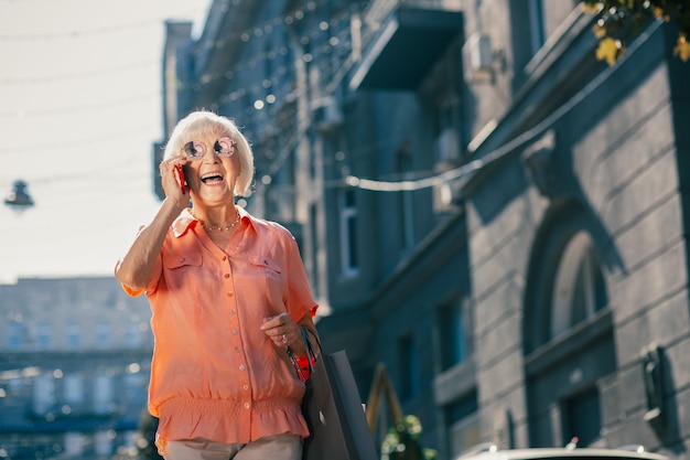 Pozytywna starsza kobieta w okrągłych okularach przeciwsłonecznych nosząca papierowe torby i uśmiechająca się z otwartymi ustami podczas rozmowy telefonicznej