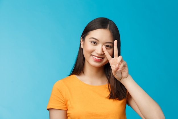 Pozytywna urocza i głupia azjatycka dziewczyna pozuje fotografię uroczo uśmiechnięta, pokazuje znak zwycięstwa pokoju przy twarzy uśmiechając się przyjazny, beztroski entuzjastyczny nastrój, stoi na niebieskim tle w żółtej koszulce.