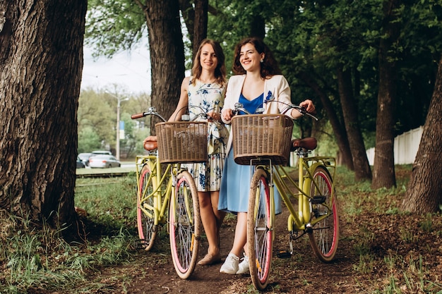 Pozytywne I Szczęśliwe Dziewczyny Chodzi Z Bicyklami Na Aleja Parku, Letni Dzień. Koleżanki, Ciesząc Się Na Spacer Ulicą Z Rowerami.