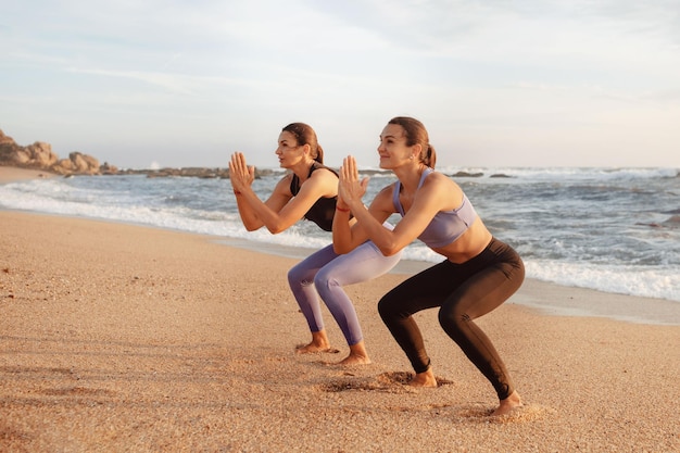 Pozytywne, ładne młode kobiety rasy kaukaskiej lubią ćwiczyć pilates razem przy przysiadach na plaży morskiej