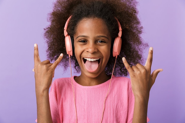 pozytywne optymistyczne szczęśliwe młode afrykańskie dziecko dziewczyna pozuje na białym tle nad fioletową ścianą, słuchając muzyki przez słuchawki pokazujące rockowy gest.