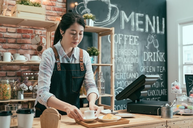 Pozytywne Uśmiechnięte Kobiece Barista Pracujący Za Drewnianym Barem. Nowoczesne Wnętrze Koncepcji Kawiarni Startowej. Młoda Dziewczyna Barman W Fartuchu Przygotować Zamówienie Klienta Z Gorącą Latte I Rogalikiem.