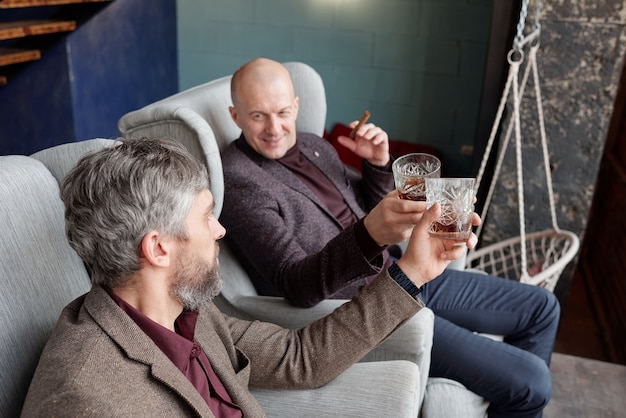 Pozytywni, bogaci panowie w średnim wieku w stylowych strojach siedzący w fotelach i relaksujący się przy whisky po pracy