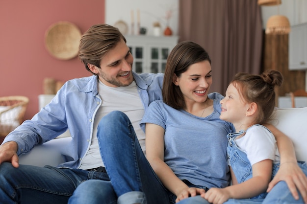 Pozytywni przyjaźni młodzi rodzice z uśmiechniętą małą córeczką siedzącą razem na kanapie, podczas relaksu w domu w weekend.