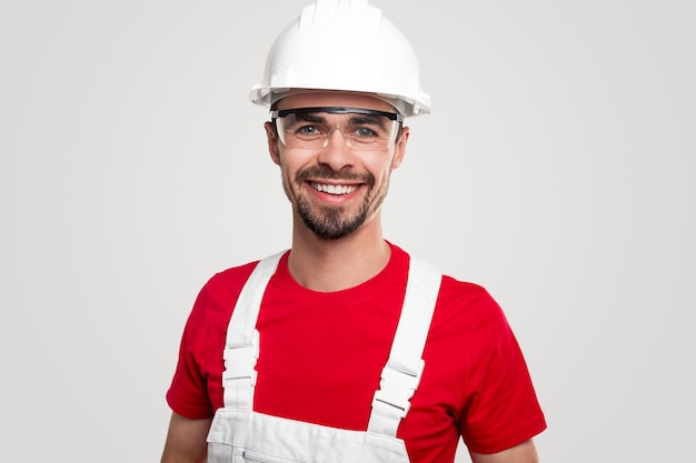Pozytywnie przyjazny brodaty pracownik płci męskiej w ogólnym i ochronnym kasku i okularach uśmiechający się i patrzący na kamerę na białym tle