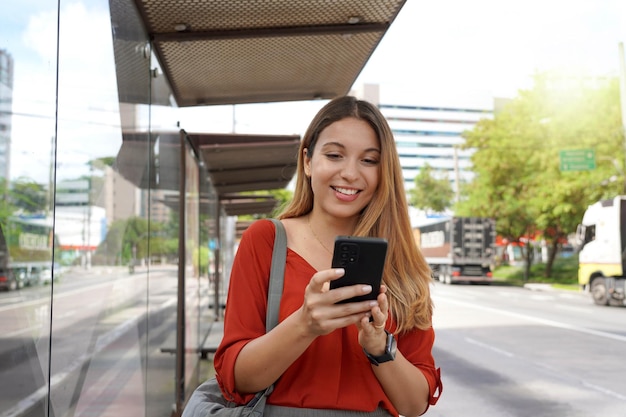 Pozytywnie uśmiechnięta dziewczyna kupująca bilet online ze smartfonem na przystanku autobusowym