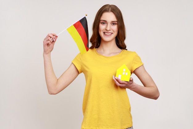 Pozytywnie uśmiechnięta nastoletnia dziewczyna w żółtej casualowej koszulce z małym papierowym domkiem i trzymająca flagę niemiec, zadowolona z zakupu zakwaterowania. Kryty studio strzał na białym tle na szarym tle.