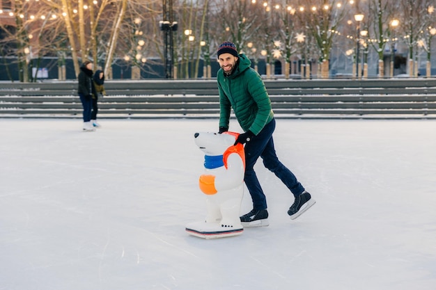 Pozytywnie uśmiechnięty atrakcyjny mężczyzna w ciepłych zimowych ubraniach i łyżwach jeździ na łyżwach używa lodowisko wygląda uroczo w kamerze spędza niedzielę na świeżym powietrzu w okresie zimowym