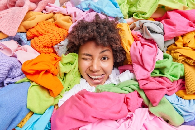 Pozytywnie zabawna Afroamerykanka otoczona kolorowymi używanymi ubraniami zebranymi do recyklingu lub darowizny czyści jej garderobę trzyma głowę przez stos wielokolorowych ubrań pokazuje język
