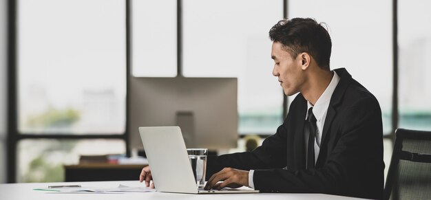 Pozytywny azjatycki mężczyzna przedsiębiorca siedzi przy stole i przegląda netbook podczas pracy nad projektem w miejscu pracy i patrząc na ekran laptopa.