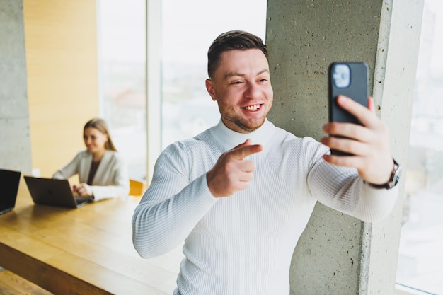 Pozytywny biznesmen w swetrze uśmiechający się patrząc na telefon stojący w nowoczesnym lekkim biurze z telefonem pracującym w nowoczesnej firmie