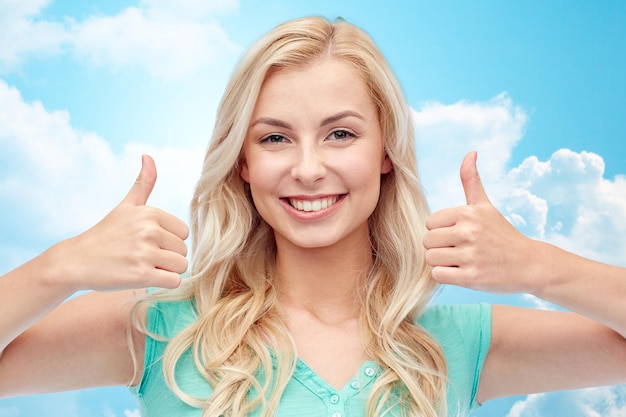 pozytywny gest i koncepcja ludzi - uśmiechnięta młoda kobieta lub nastolatka pokazująca kciuki obiema rękami na tle błękitnego nieba i chmur