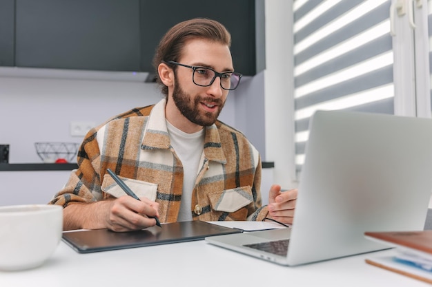 Pozytywny mężczyzna w okularach pracujący z tabletem graficznym w domu za laptopem