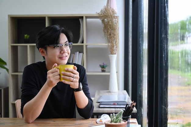 Pozytywny młody azjatycki człowiek trzyma filiżankę kawy i patrząc przez okno.