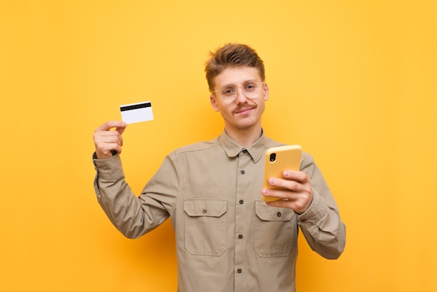 Pozytywny młody człowiek trzyma kartę kredytową i smartfon z okularami i wąsami