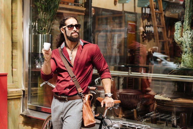 Zdjęcie pozytywny stylowy mężczyzna stojący w pobliżu roweru, ciesząc się kawą